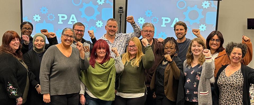 Photo de groupe de l'équipe de négociation de la PA, debout, les poings levés en signe de solidarité, devant une toile de fond sur laquelle est inscrit « PA ».