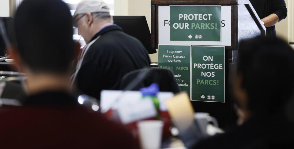 Photo de conférence de Parcs Canada Des participantes et participants assistent à une rencontre en appui au personnel de Parcs Canada, avec des affiches « Protect Our Parks » et « On protège nos parcs » exposées sur le lutrin.