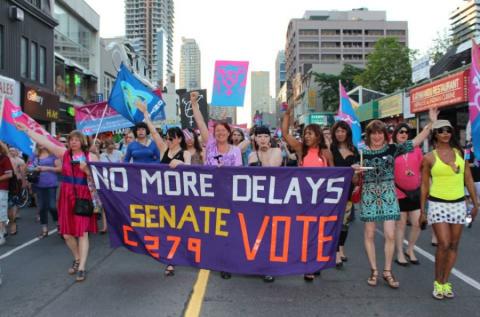 Foule colorée agitant des drapeaux et une banière critiquant les retards dans l’adoption de C-279 à la marche mondiale pour la Fierté à Toronto