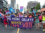 Foule colorée agitant des drapeaux et une banière critiquant les retards dans l’adoption de C-279 à la marche mondiale pour la Fierté à Toronto